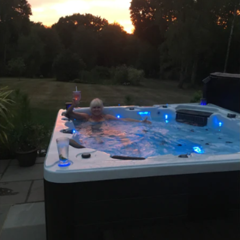 A lifestyle shot of the Vancouver 65-Jet 6-Person Hot Tub that has a lady in there raising a glass with a big smile on her face. You can see behind the hottub is a big garden with an open grass area surrounded by trees and bushes. 